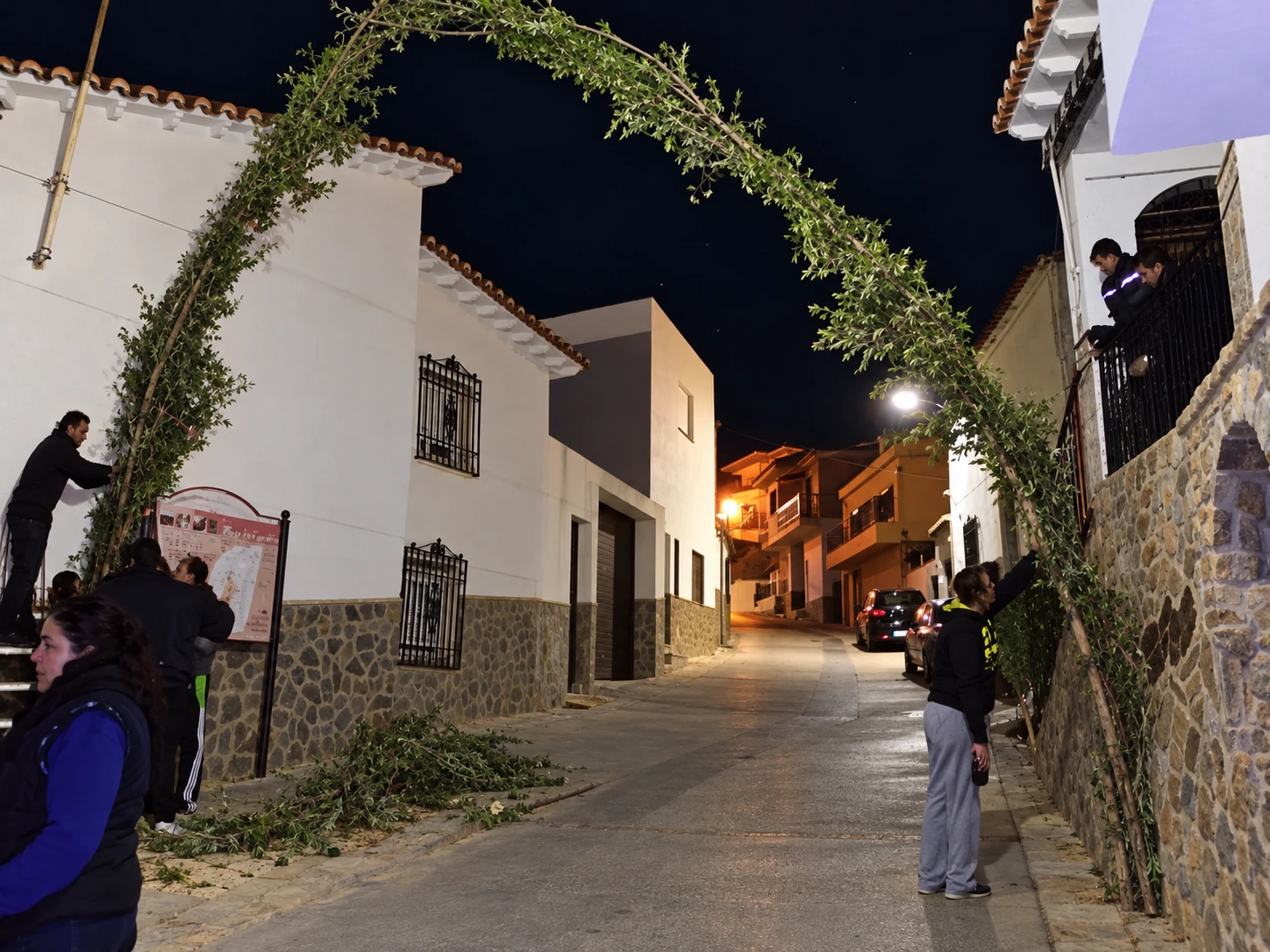 Vecinos sujetan y preparan un arco de ramas entre dos fachadas durante la noche
